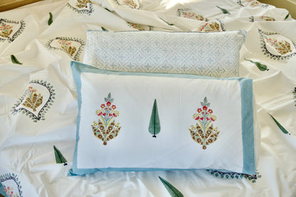 Hand block printed organic cotton bedsheet with colourful floral motifs, green cypress trees, and blue-bordered pillowcases, paired with a geometric patterned pillow.