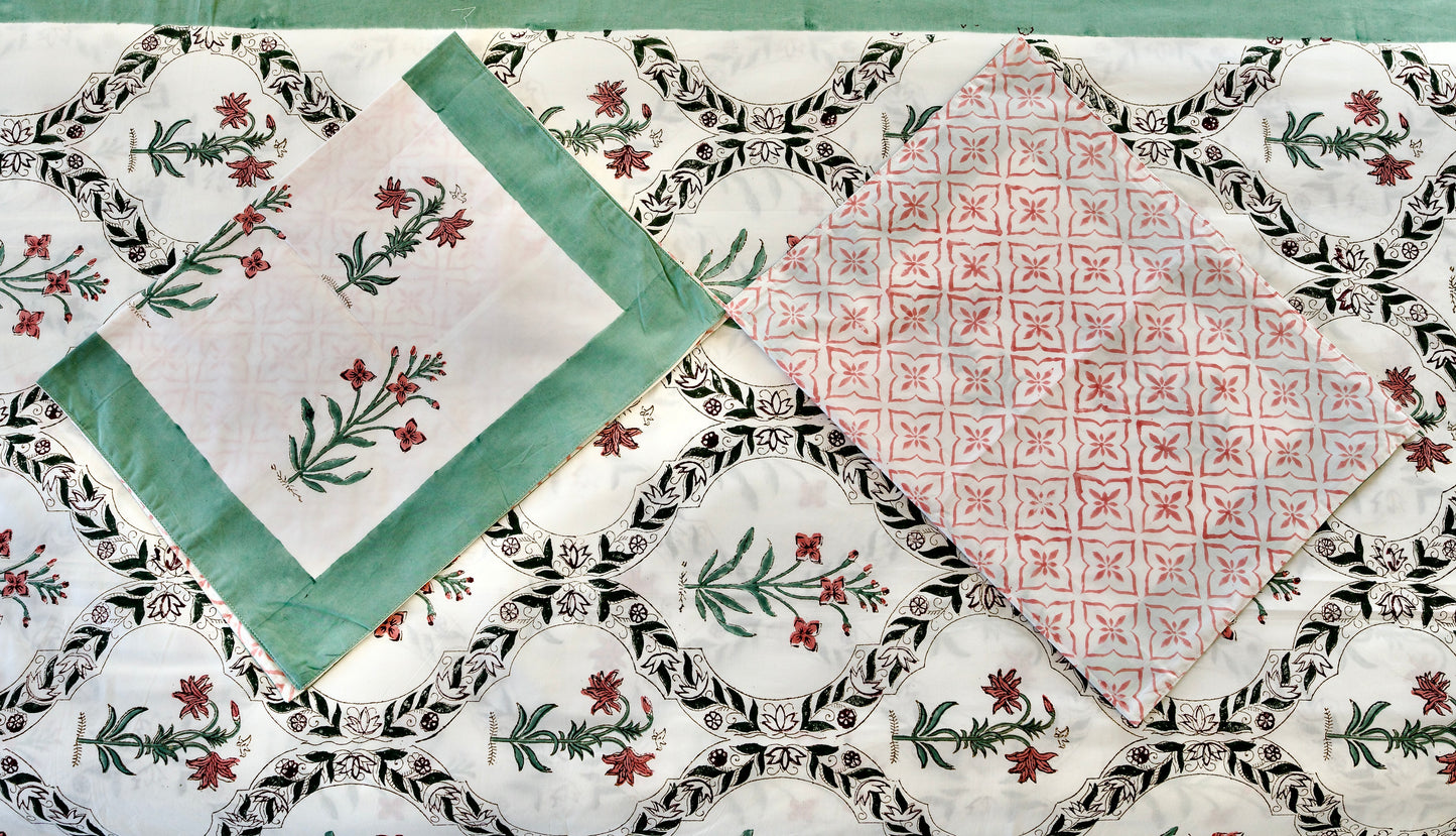 Vine Mosaic hand block printed organic cotton bedsheet with red florals and green stems framed in black vine arches on a white base, paired with one green-bordered floral pillowcase and one red geometric patterned pillowcase.