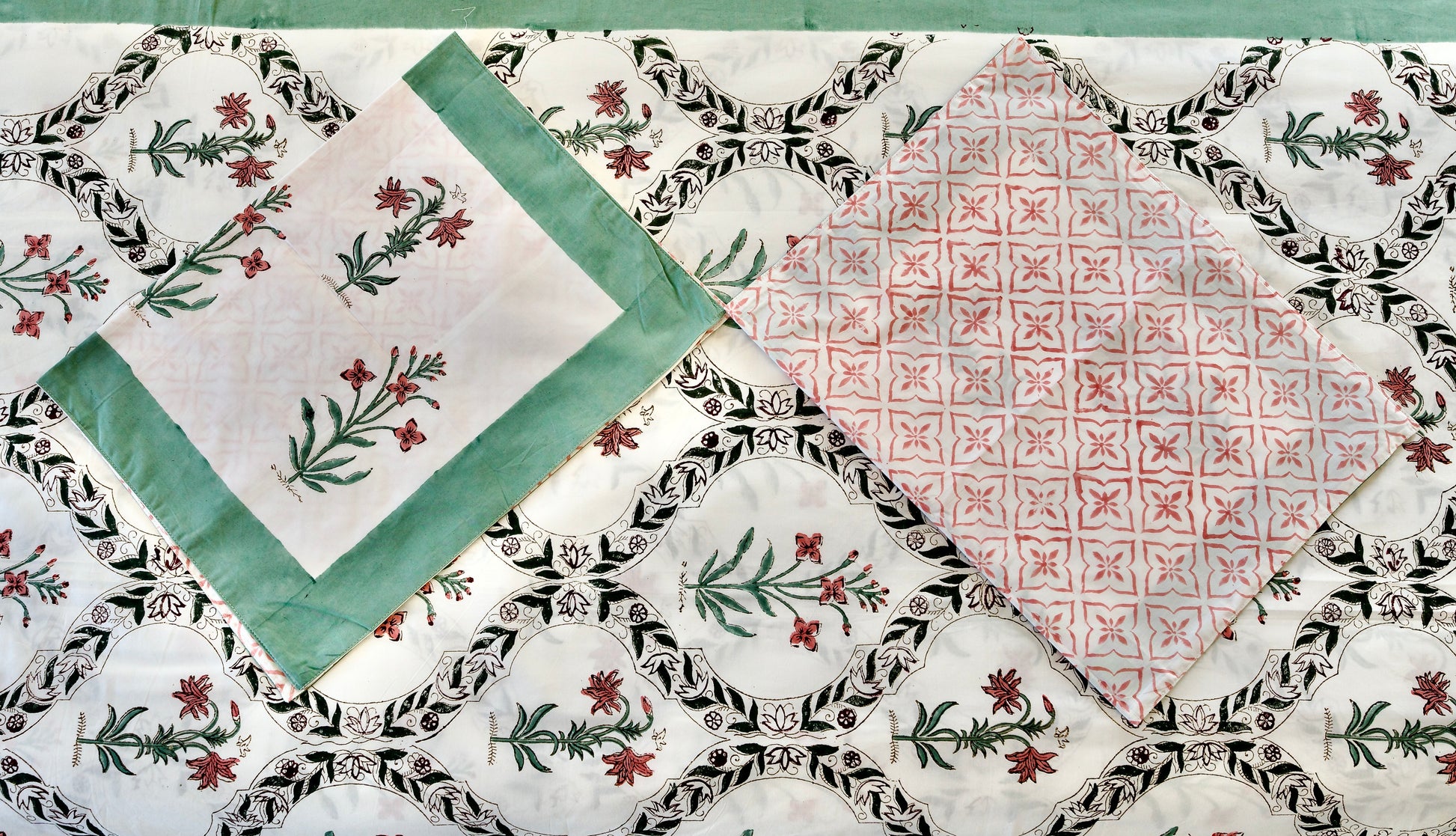 Vine Mosaic hand block printed organic cotton bedsheet with red florals and green stems framed in black vine arches on a white base, paired with one green-bordered floral pillowcase and one red geometric patterned pillowcase.